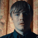 rayne, a man with a brown bowlcut and blue eyes wearing a black dress shirt